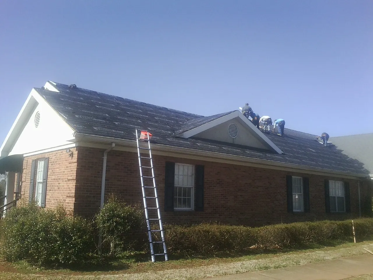 Crew tearing shingles off a brick ranch roof with a ladder set — Emergency Roof Repair work by our crew in Fabius