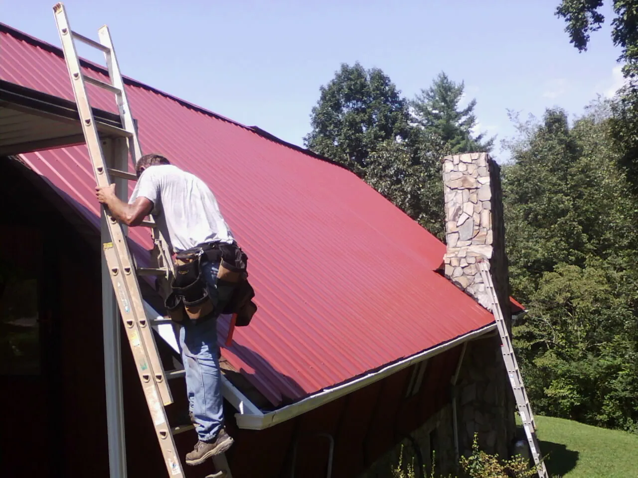 Roofer on a ladder beside a red metal roof and stone chimney — Soffit & Fascia Repair by Roofing Brothers in Fabius